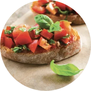 Une photo circulaire d'une tartine de pain sur laquelle est posée des tomates coupées en petits morceaux. Des feuilles de basilic sont parsemées sur le tout.